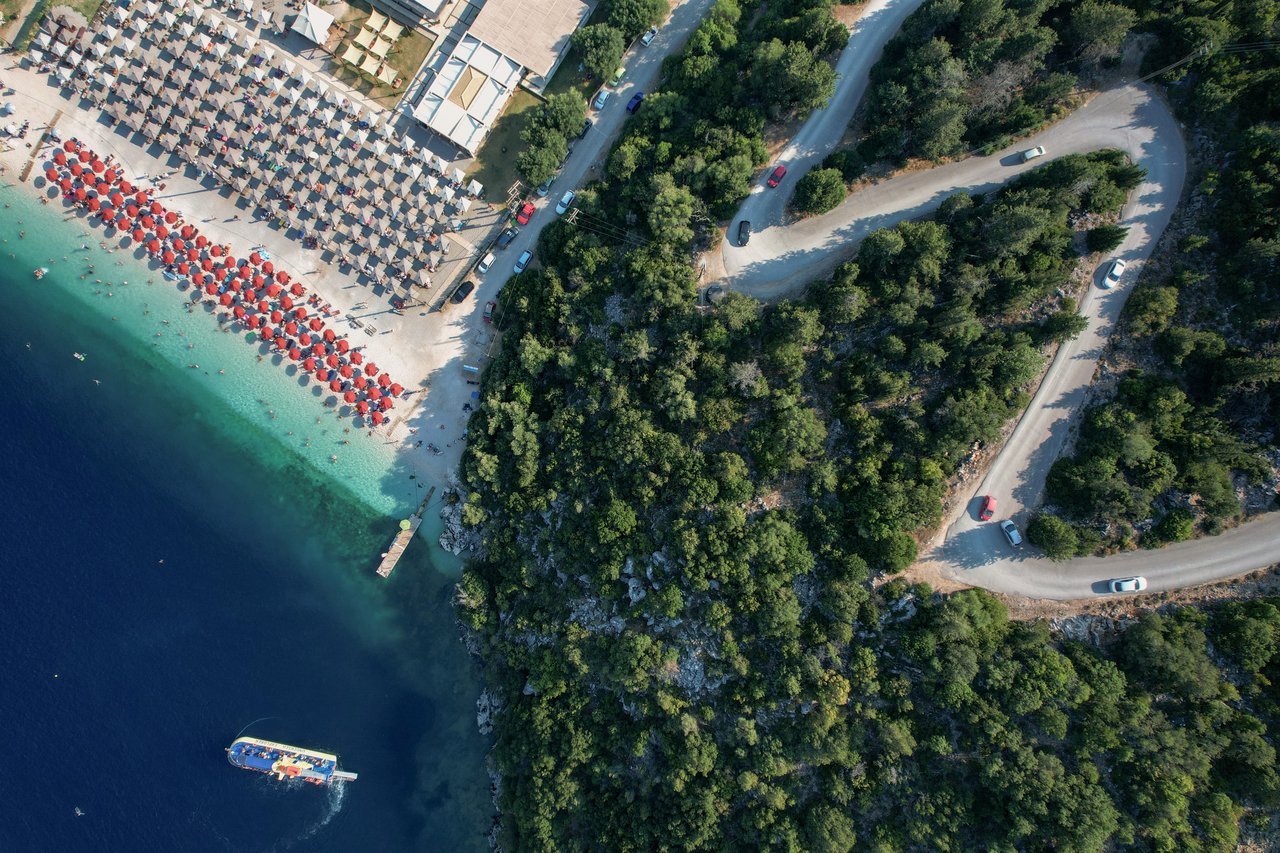 Seen from above, a winding road through the trees down to a beach with red umbrellas in Greece.