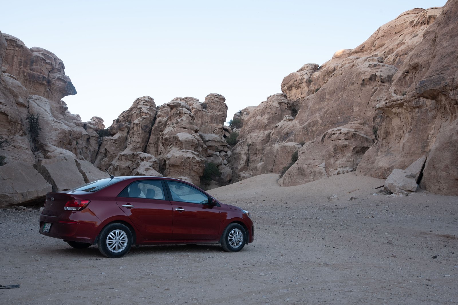 A red sedan parked on a sandy track below the sandstone cliffs of Little Petra, Jordan.