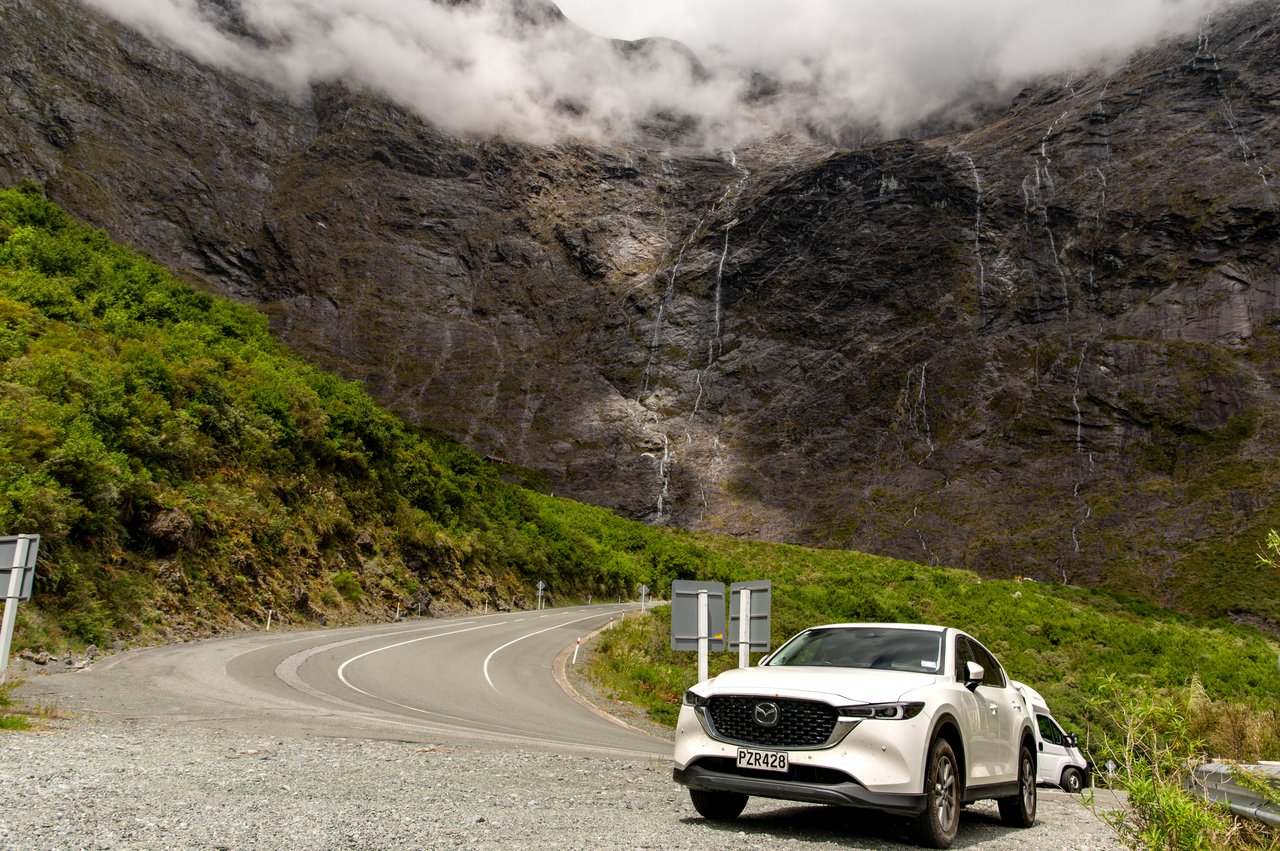 A white Mazda SUV parked beside the winding road to Milford Sound, under waterfalls running down a steep mountain wall, just past the Homer Tunnel in New Zealand.