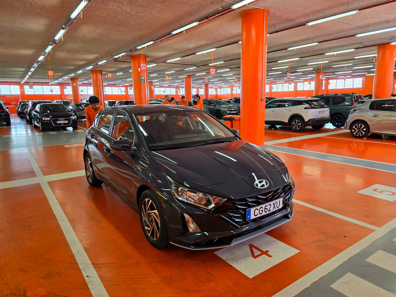 A dark gray Hyundai hatchback in bay 4 of a rental car garage in Lisbon, with orange pillars and staff in orange behind it.